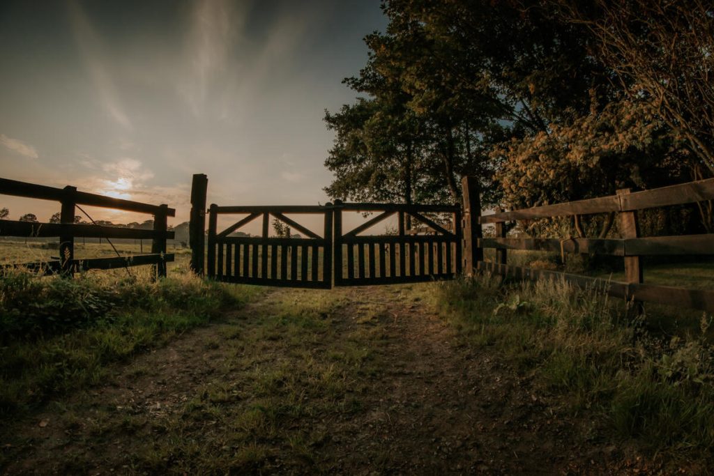 A timber farm gate closed across a track at dusk, with the sun low behind the trees